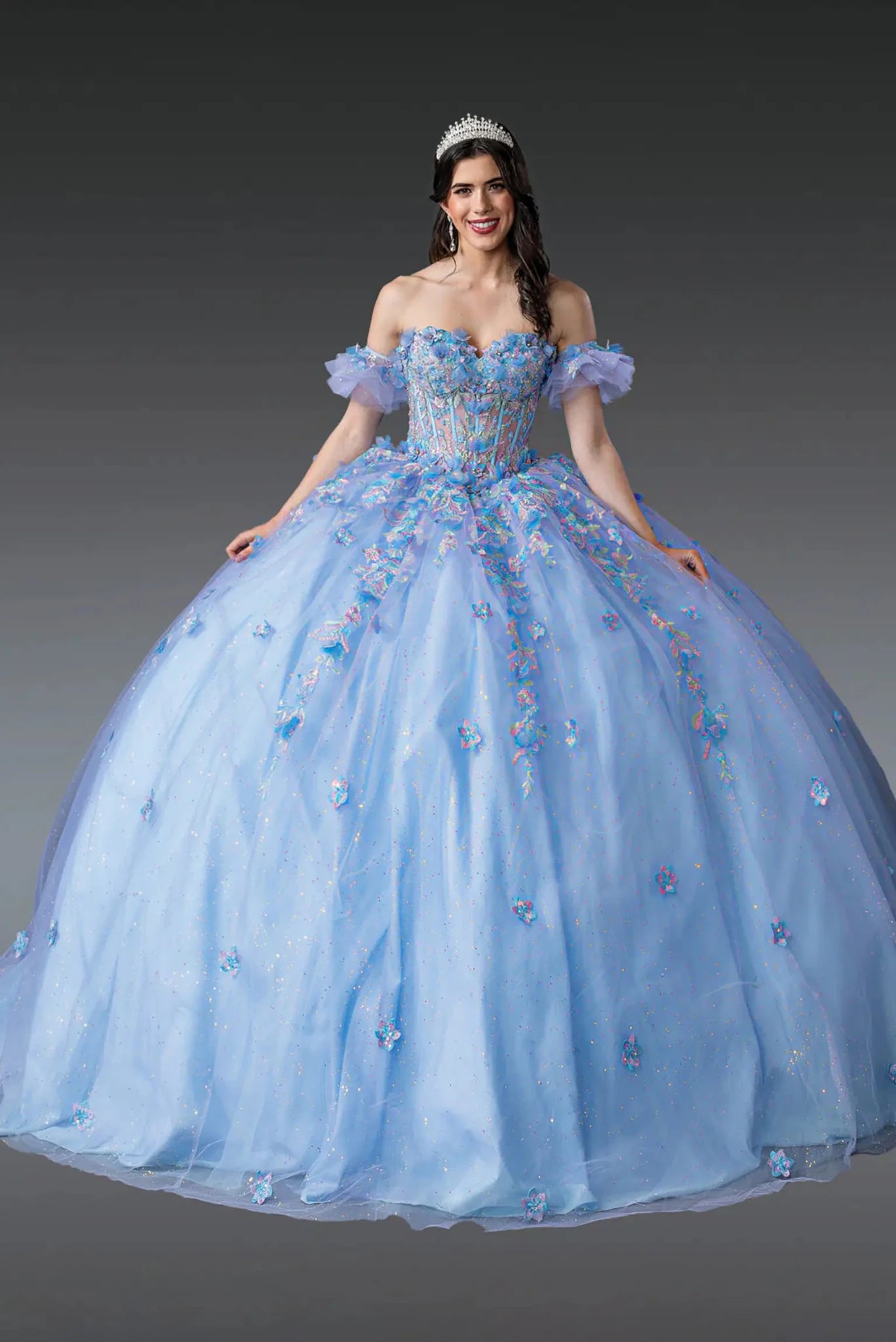 Woman standing front-facing in Bahama blue-lilac quinceañera ball gown with off-shoulder sleeves, beaded bodice, floral appliqués, and voluminous full tulle skirt with delicate embellishments
