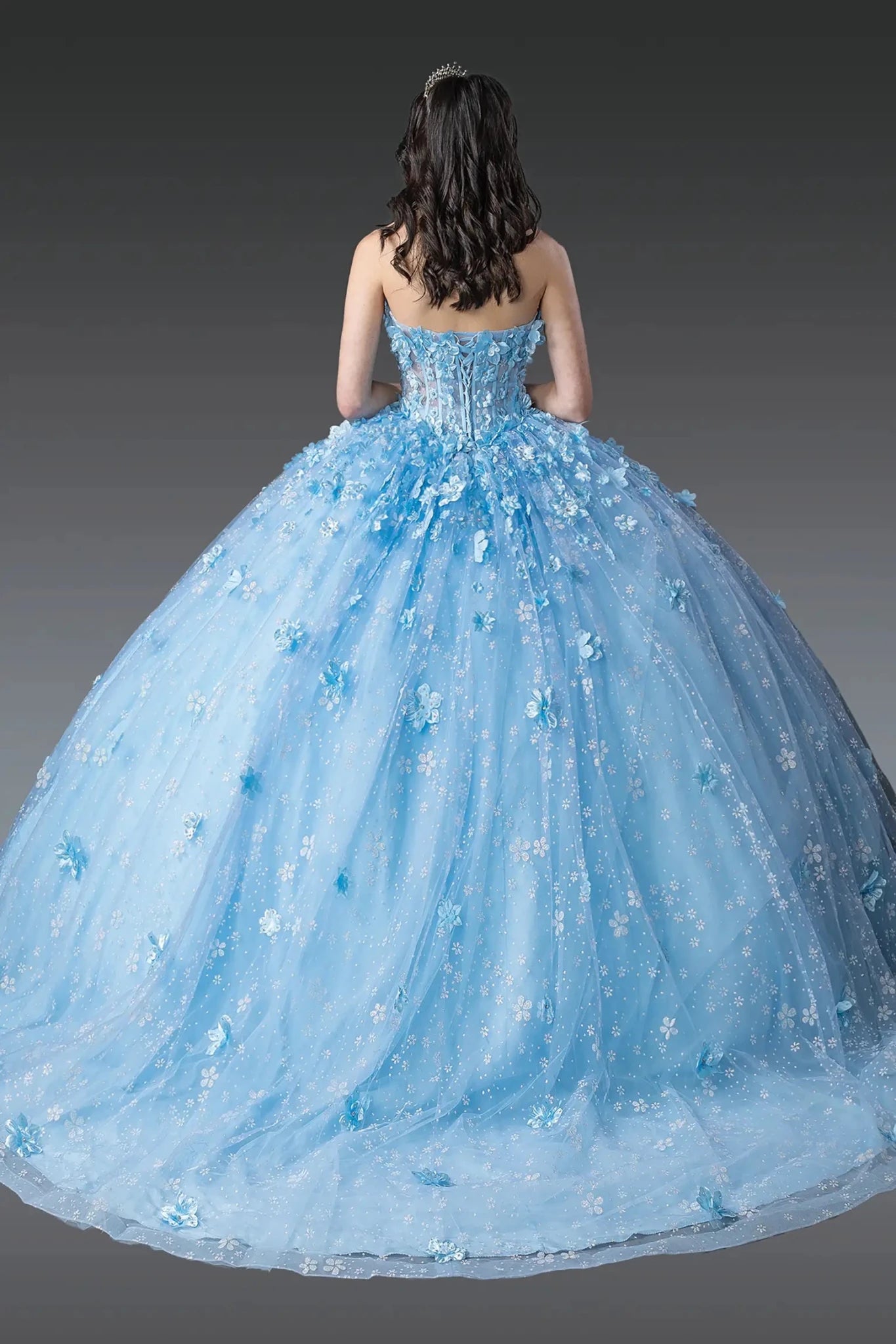 Woman standing back-facing in Bahama blue quinceañera ball gown with lace-up beaded bodice, off-shoulder sleeves, floral embellishments, and flowing glitter tulle skirt with train