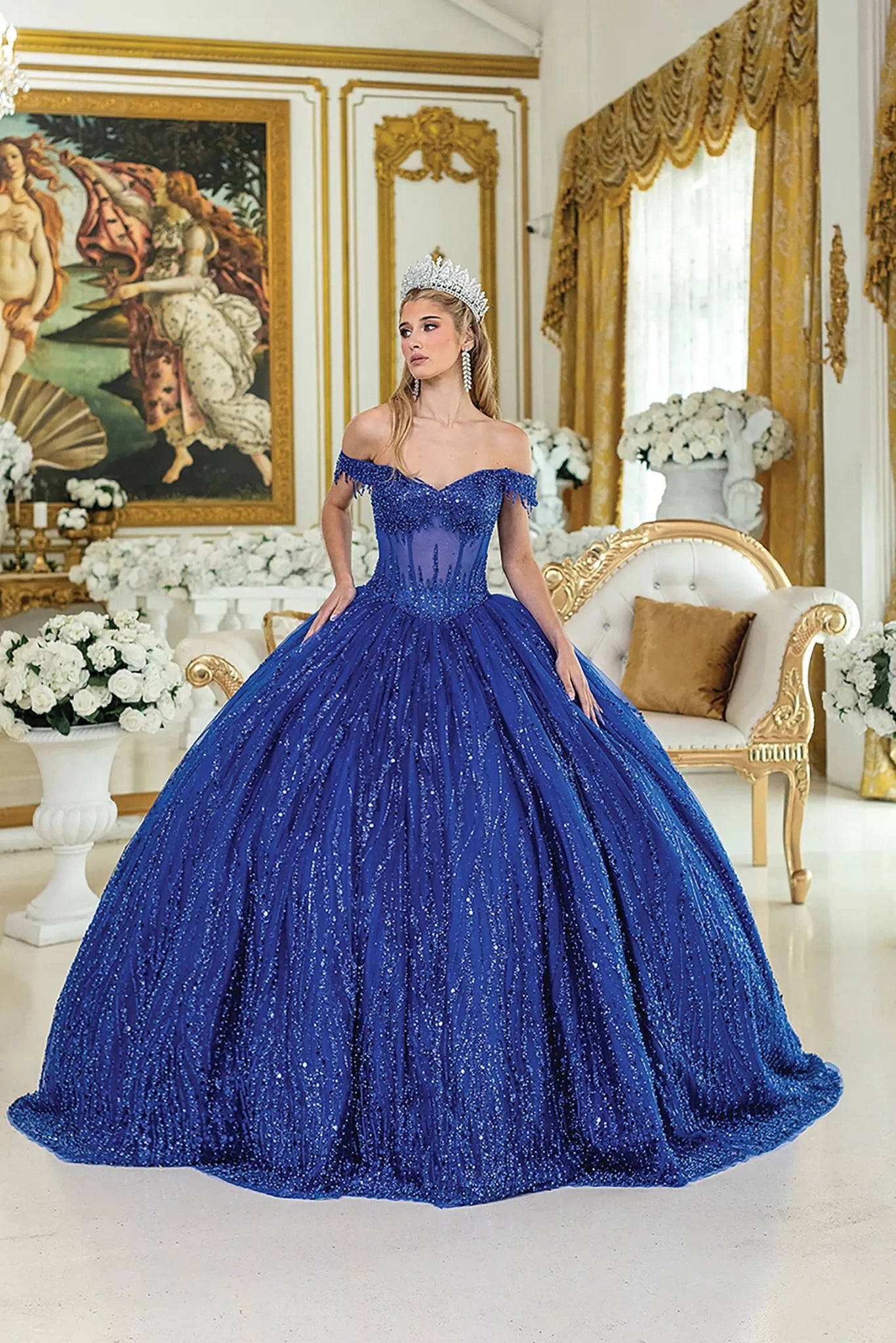 Woman standing front-facing in royal blue quinceañera ball gown with off-shoulder sleeves, sequined overlay, beaded bodice, and layered glittering skirt