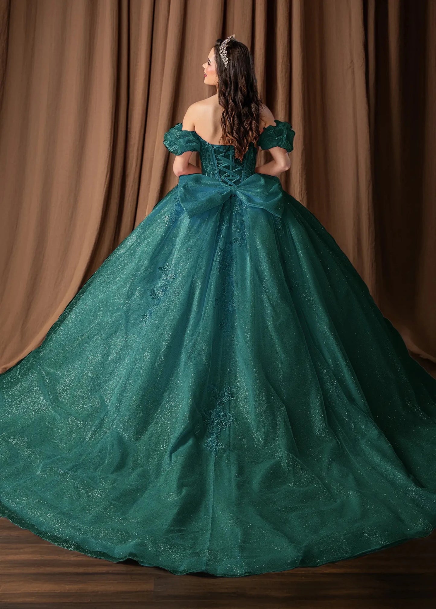 Woman standing back-facing in hunter green quinceañera ball gown with off-shoulder ruffled sleeves, embroidered bodice, and shimmering layered skirt