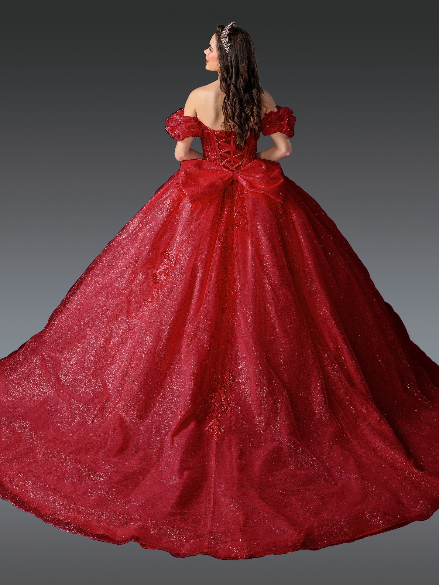 Woman standing back-facing in red quinceañera ball gown with off-shoulder sleeves, lace-up embroidered bodice, and flowing layered skirt with train