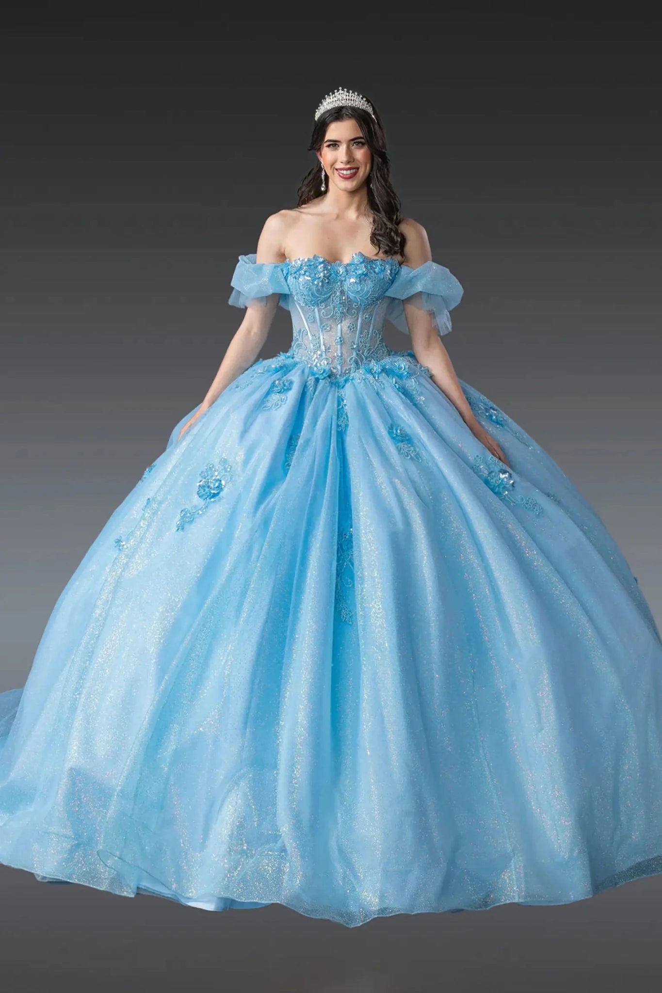Woman standing front-facing in Bahama blue quinceañera ball gown with off-shoulder sleeves, beaded corset bodice, floral embellishments, and voluminous layered tulle skirt with glitter accents