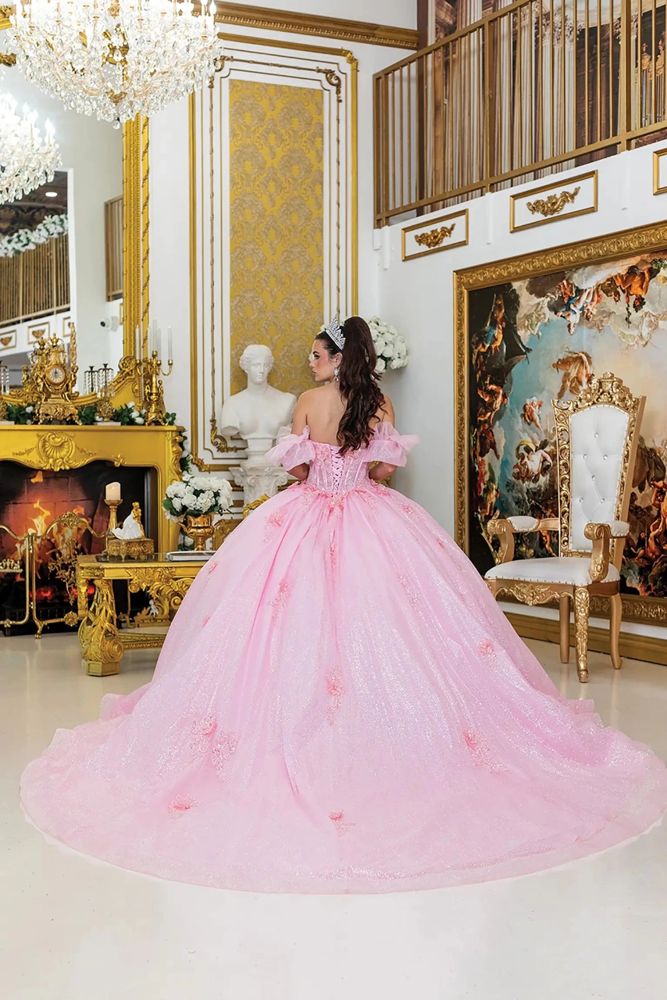Woman standing back-facing in pink quinceañera ball gown with lace-up bodice, embroidered floral details, sheer overlay, and flowing layered skirt with train