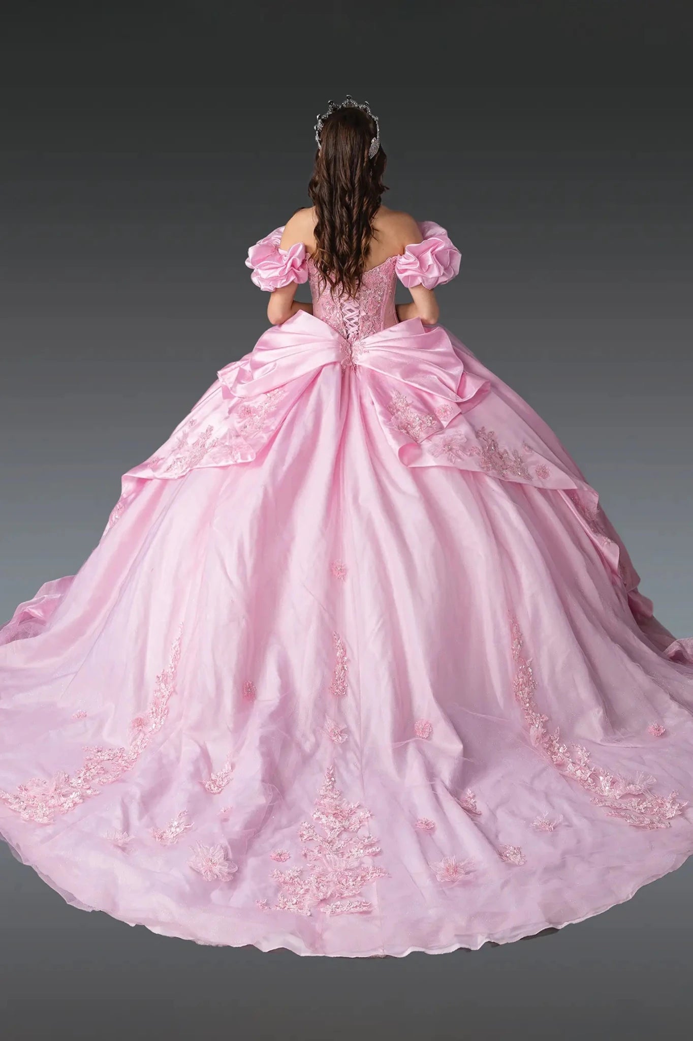 Woman standing back-facing in pink quinceañera ball gown with lace-up corset bodice, puff sleeves, floral embroidery, and flowing layered tulle skirt with train