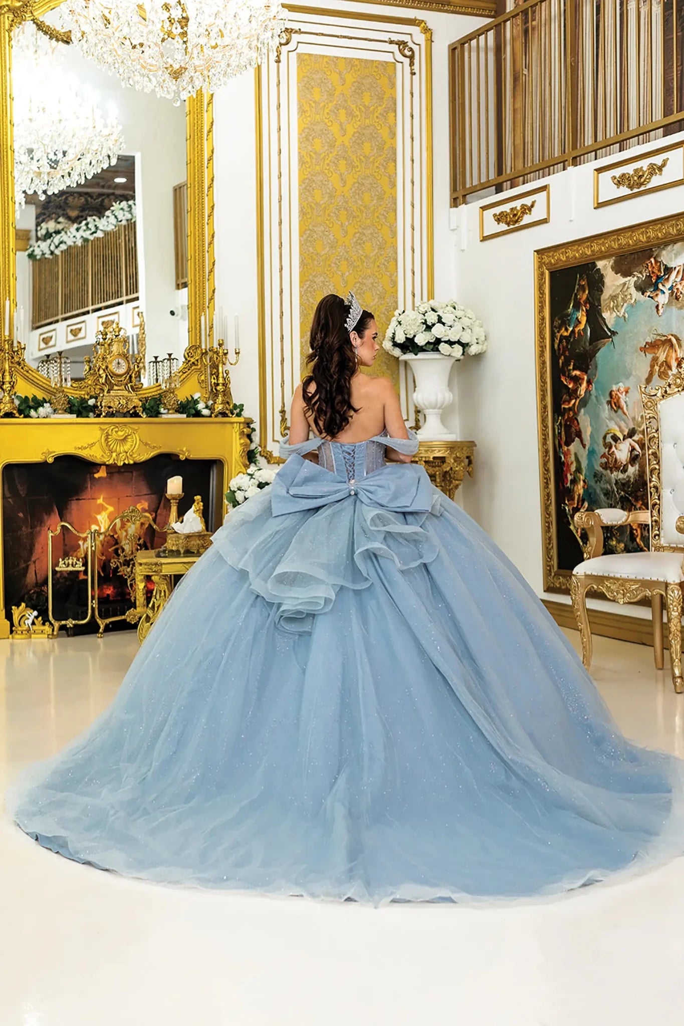 Woman standing back-facing in pink strapless quinceañera ball gown with lace-up sheer corset bodice, off-shoulder sleeves, and flowing sparkling tulle skirt with train