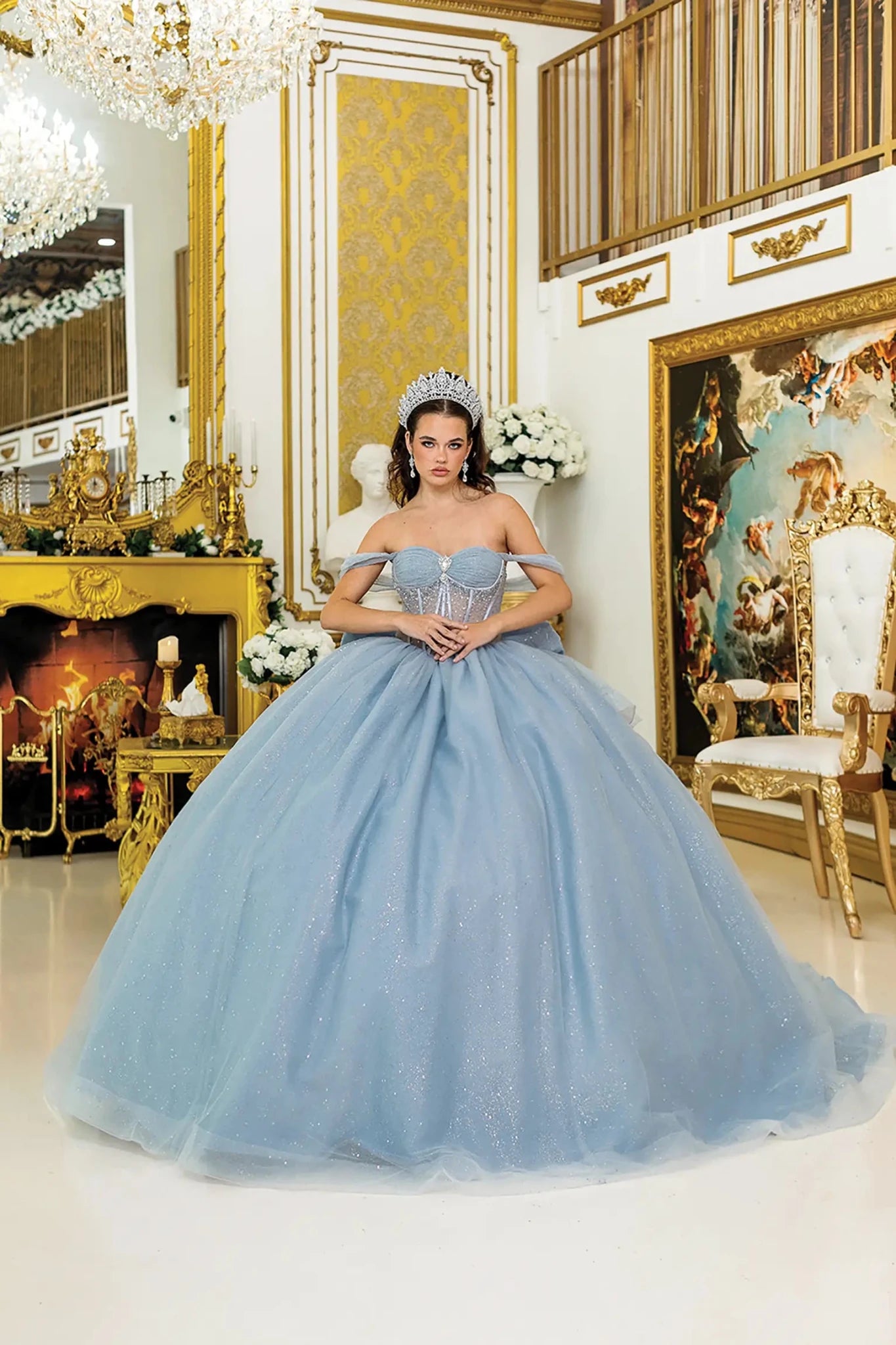 Woman standing front-facing in dusty blue strapless quinceañera dress with off-shoulder sleeves, sheer corset bodice, and sparkling voluminous ball gown skirt
