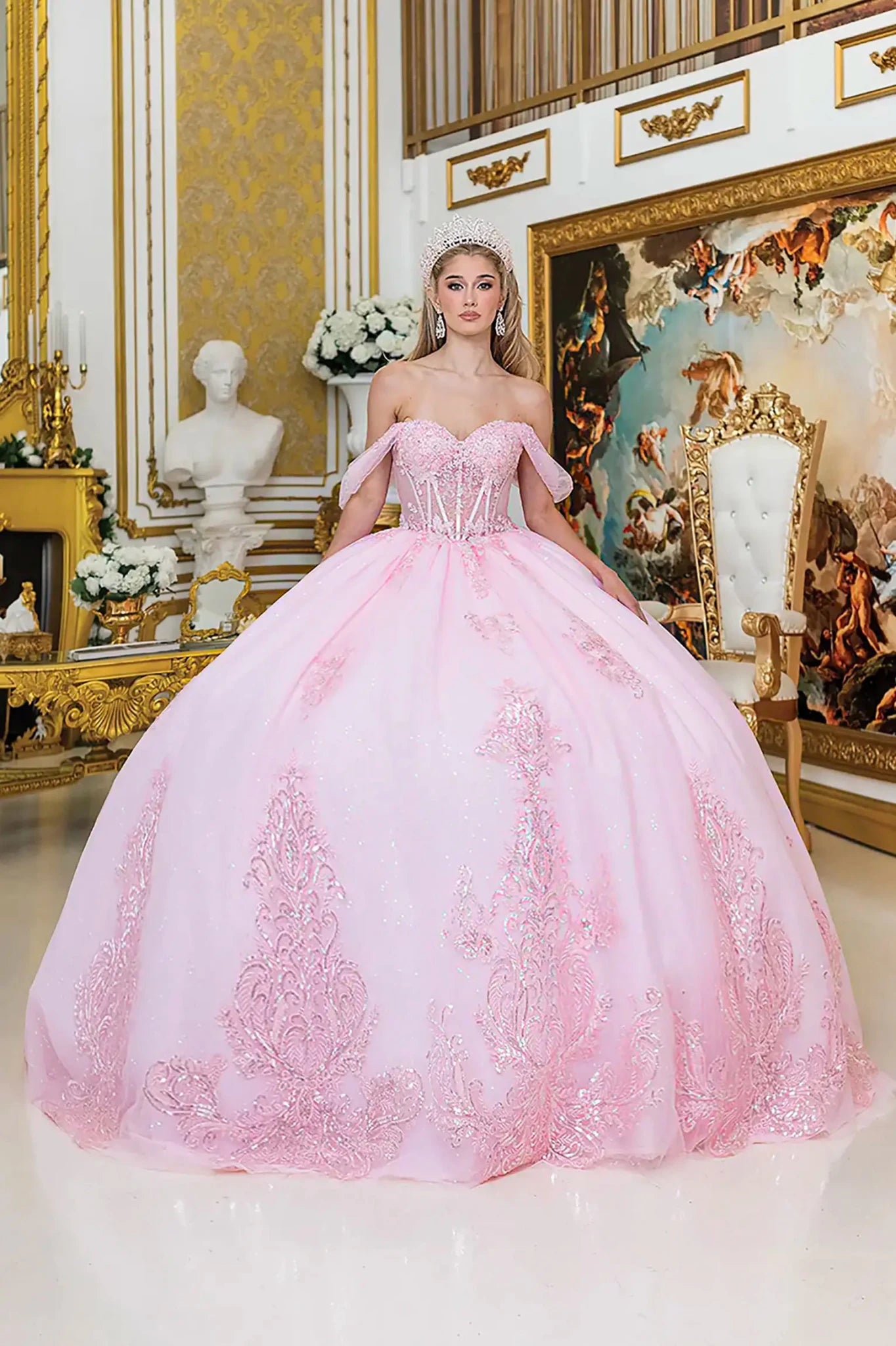 Woman in a pink ball gown with off shoulder standing in an ornate room with gold accents and decorative elements.