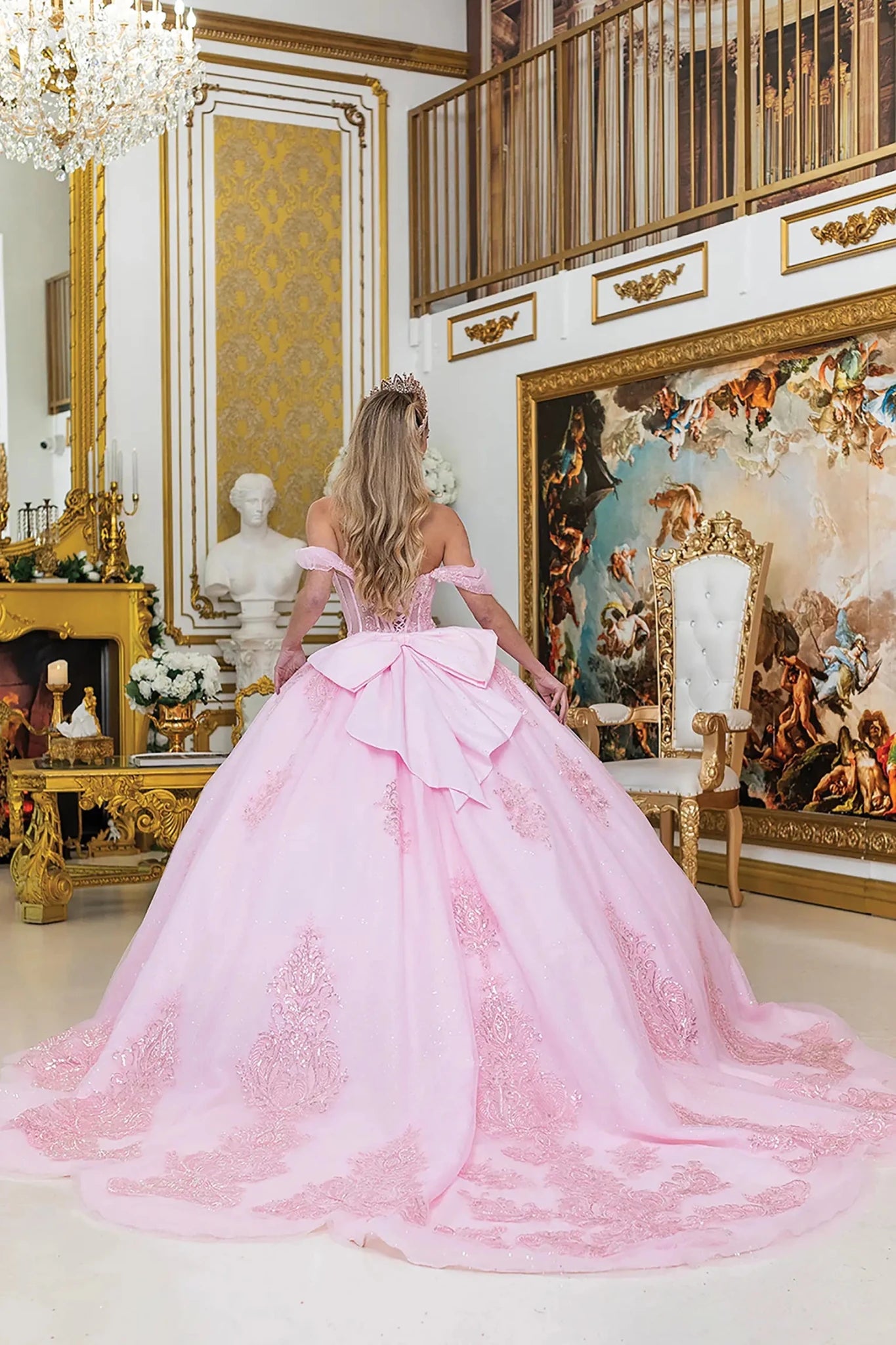 Woman in a pink ball gown with big back bow and long train standing in an ornate room with gold accents and classical art.