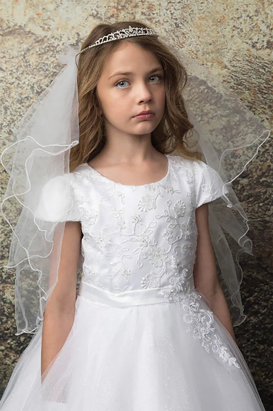 Young girl in a white dress with lace details and a veil against a textured wall.