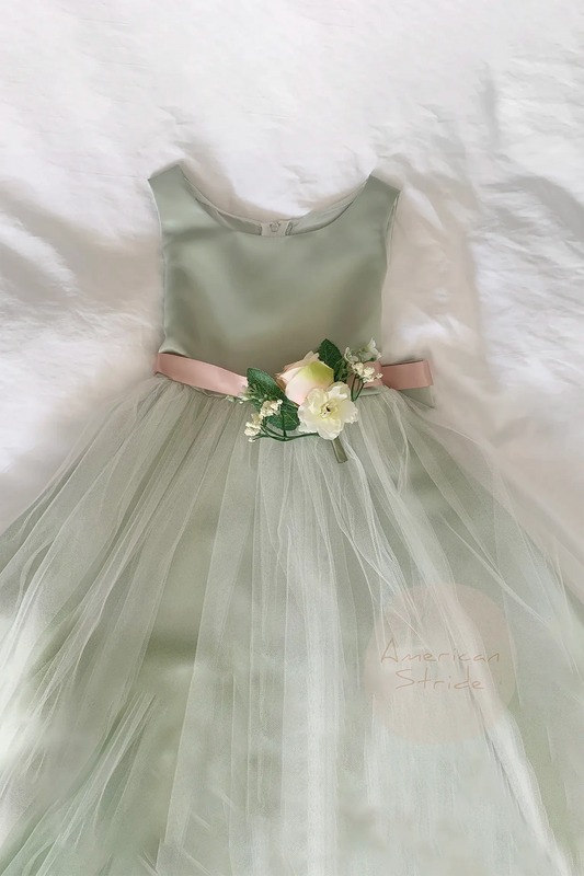 sage flower girl dress with a floral sash on a white fluffy surfacej. top is sage satin and the skirt is tulle.