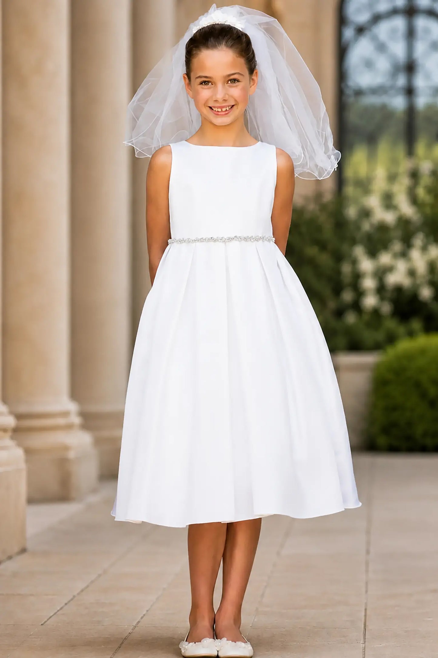 Young girl in a white dress and pearl/rhinestone belt with a veil standing against a wooden wall.