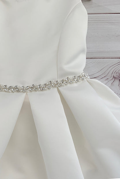 White satin flower girl dress with a decorative belt on a wooden surface.