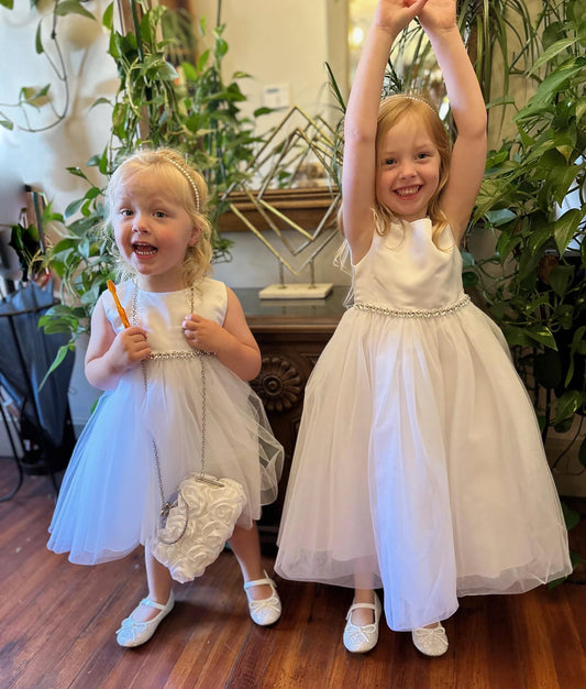 Two young girls in white dresses standing indoors with plants in the background