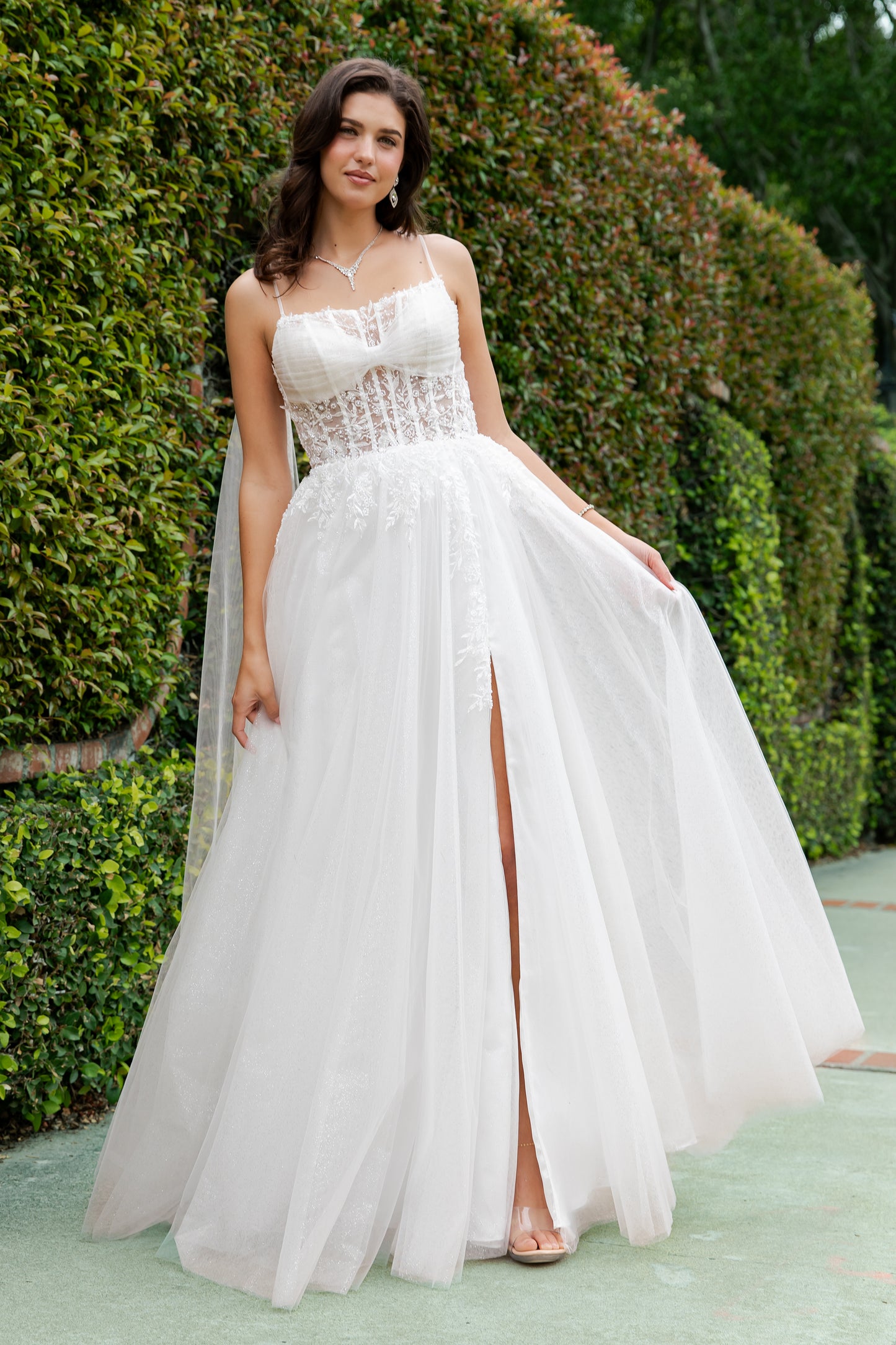 Woman in a white strapless gown with a high slit standing outdoors against greenery.