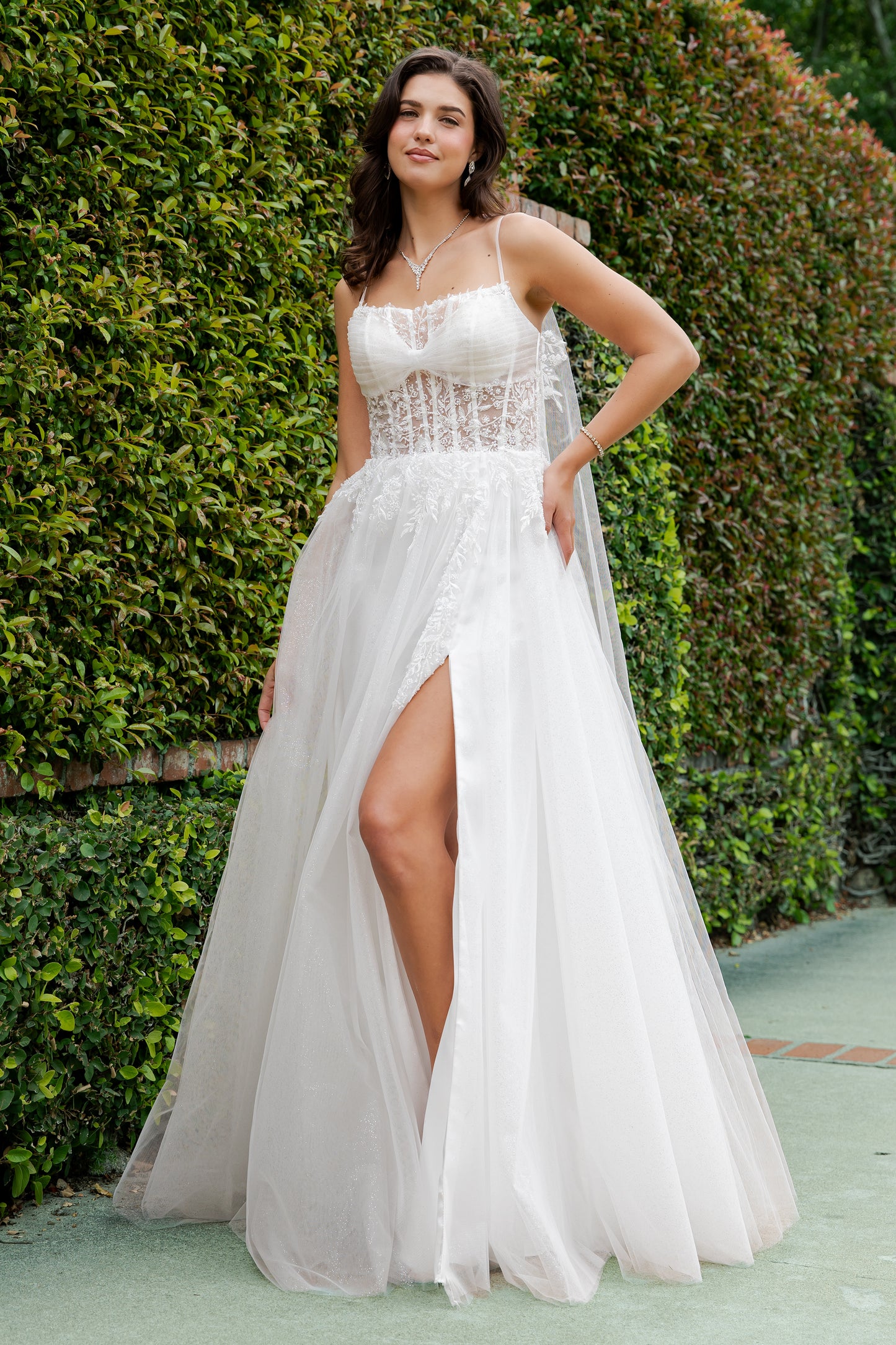 Woman in a white dress with a high slit standing in front of green hedges.