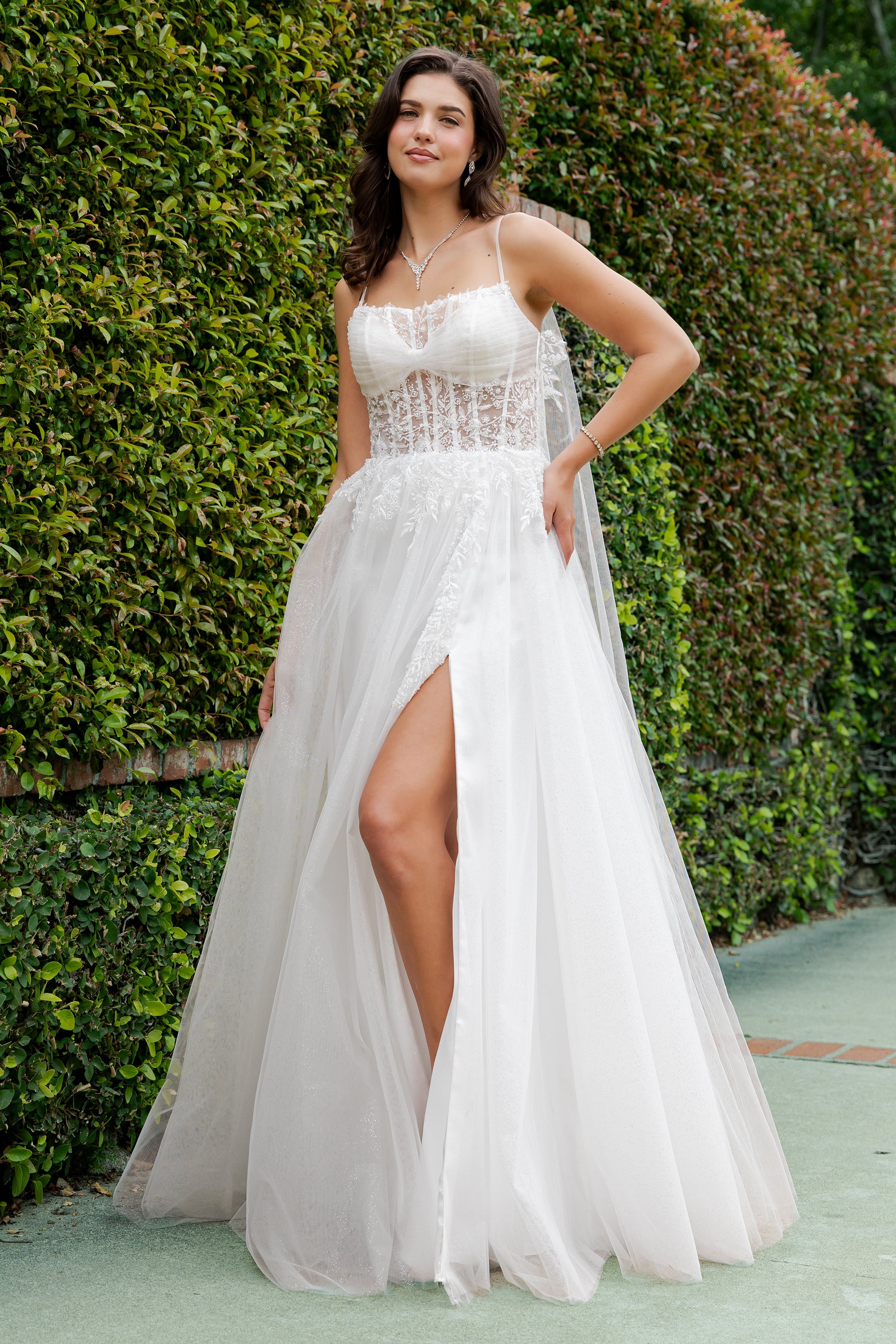 Woman in a white dress with a high slit standing in front of green hedges.