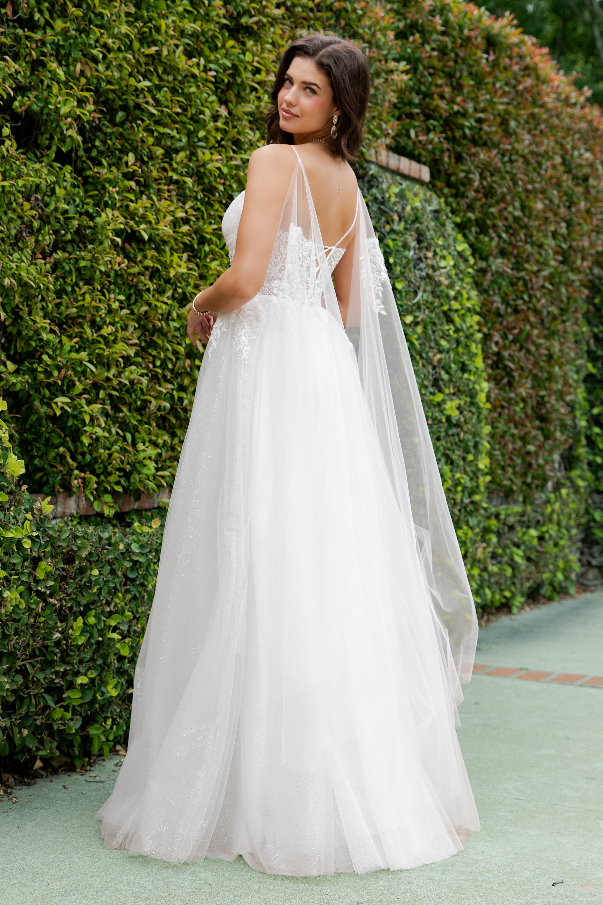 Woman in a white wedding dress with a cape standing in front of green bushes.
