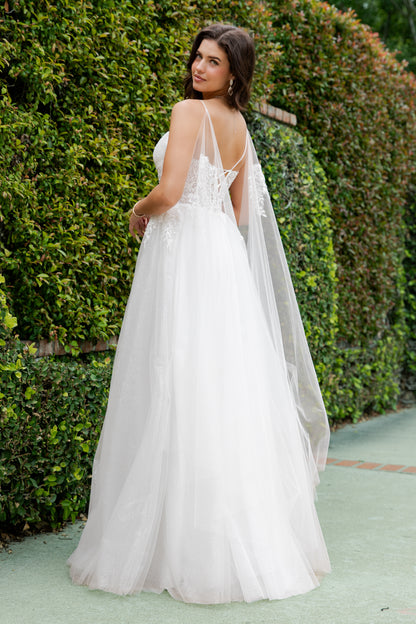 Woman in a white wedding dress with a cape standing in front of green bushes.