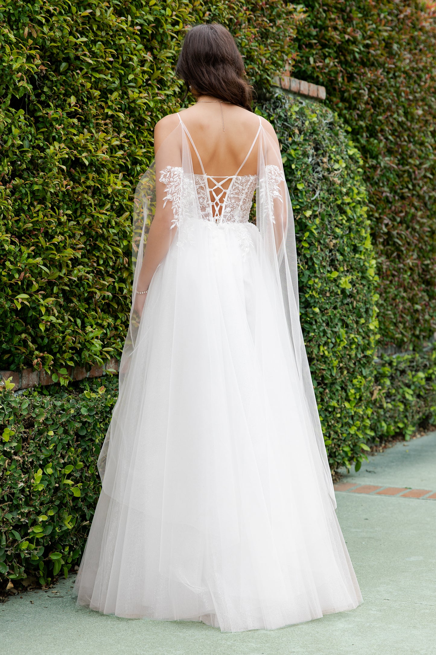 Woman wearing a white wedding dress with a lace back and sheer sleeves against a green hedge.