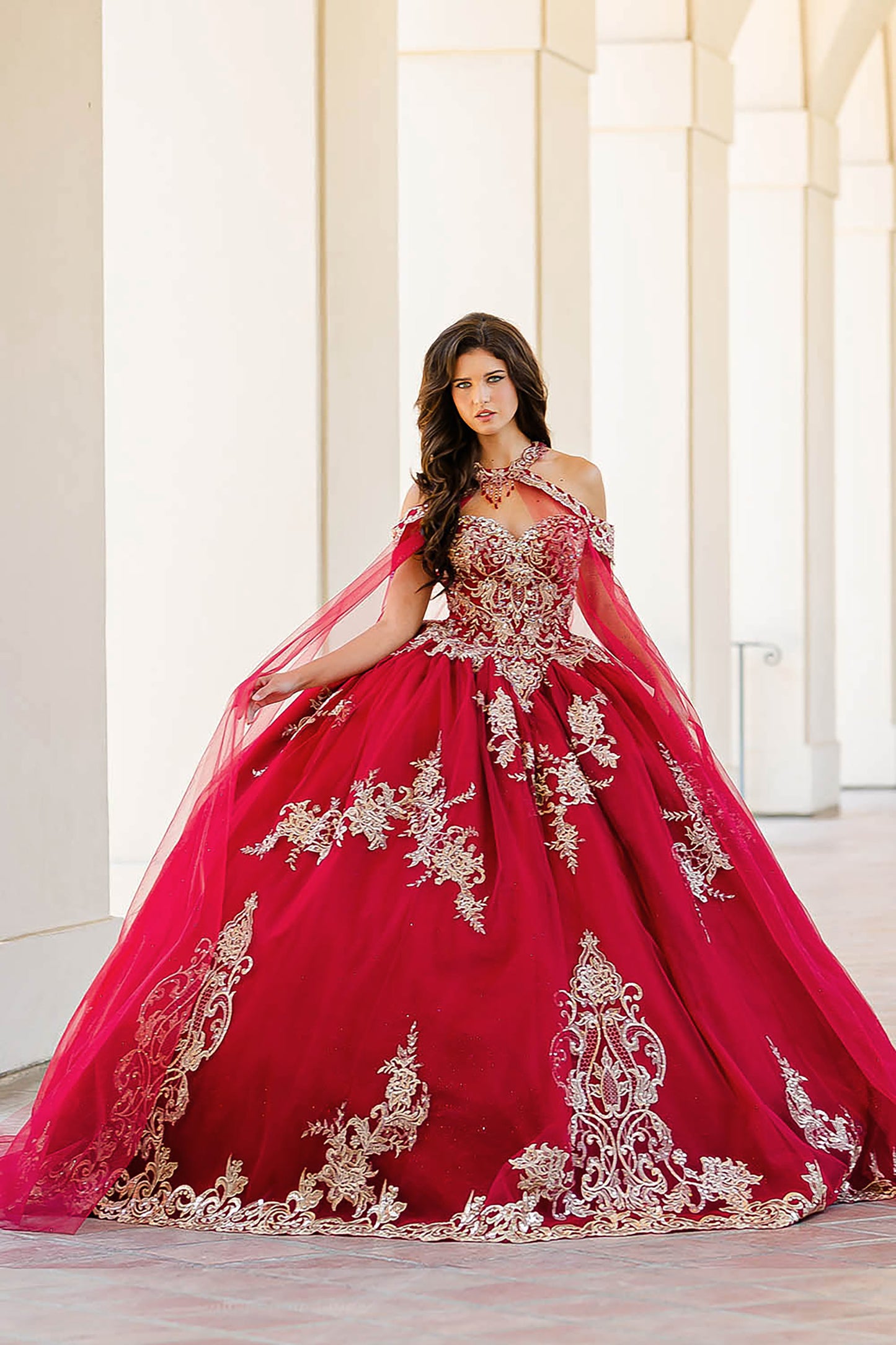 Quinceanera | Wine with Gold Embroidered Lace with Stunning Cape Quince Ballgown dress - front view of woman wearing dress standing in front of pillars