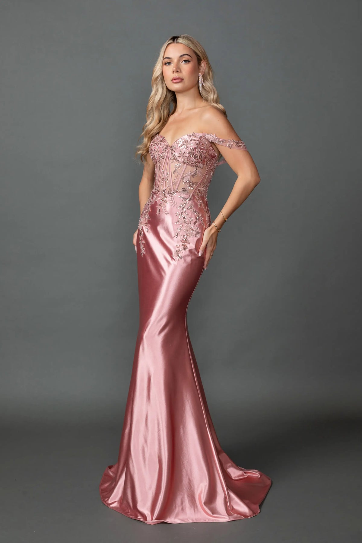 Showing beaded corset detailing above its satin skirt, this Rose Gold prom dress is shown in full view.
