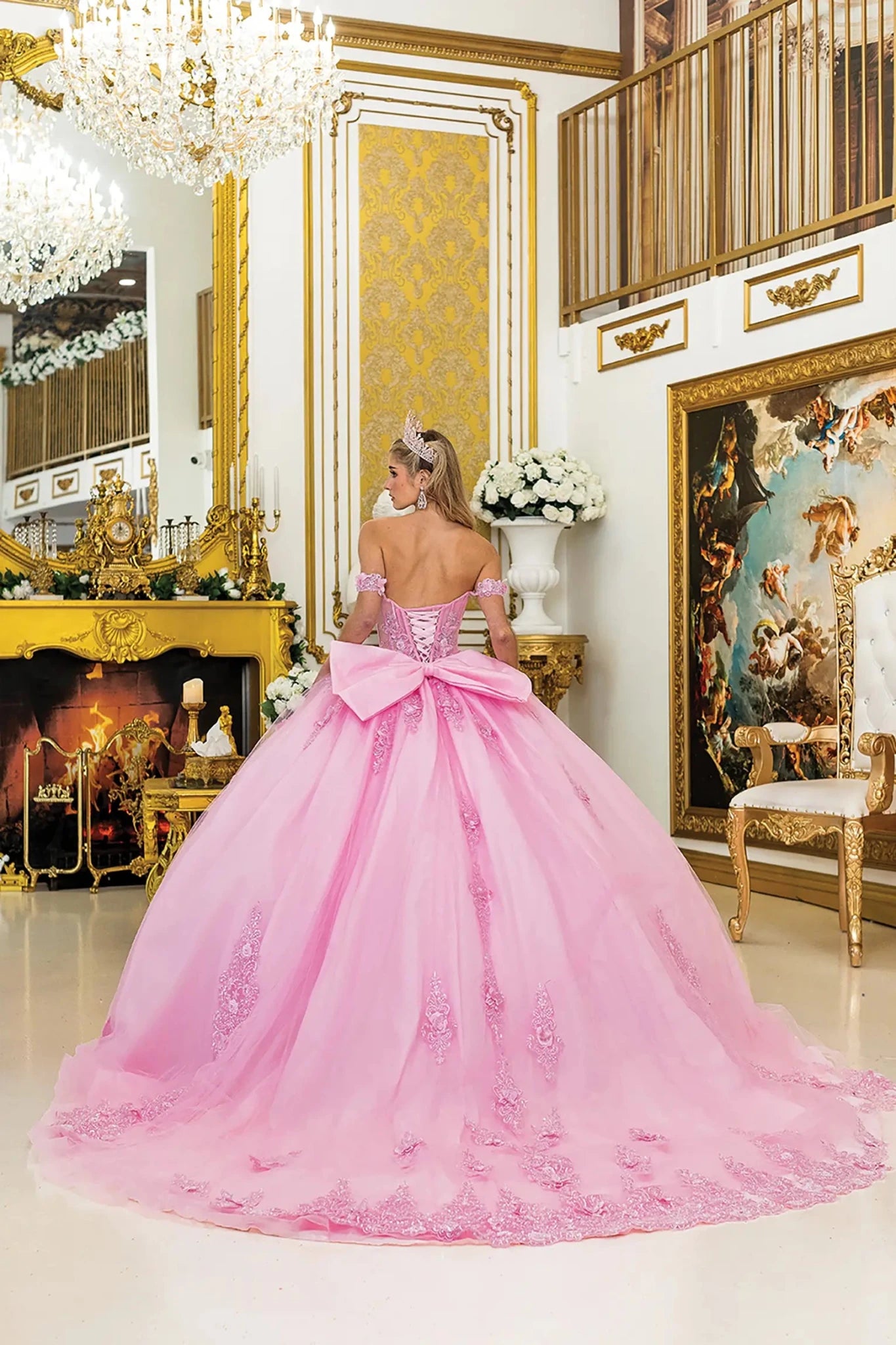 Woman standing back-facing in pink quinceañera ball gown with off-shoulder floral appliqué sleeves, lace-up sheer corset bodice, embroidered skirt accents, and flowing train