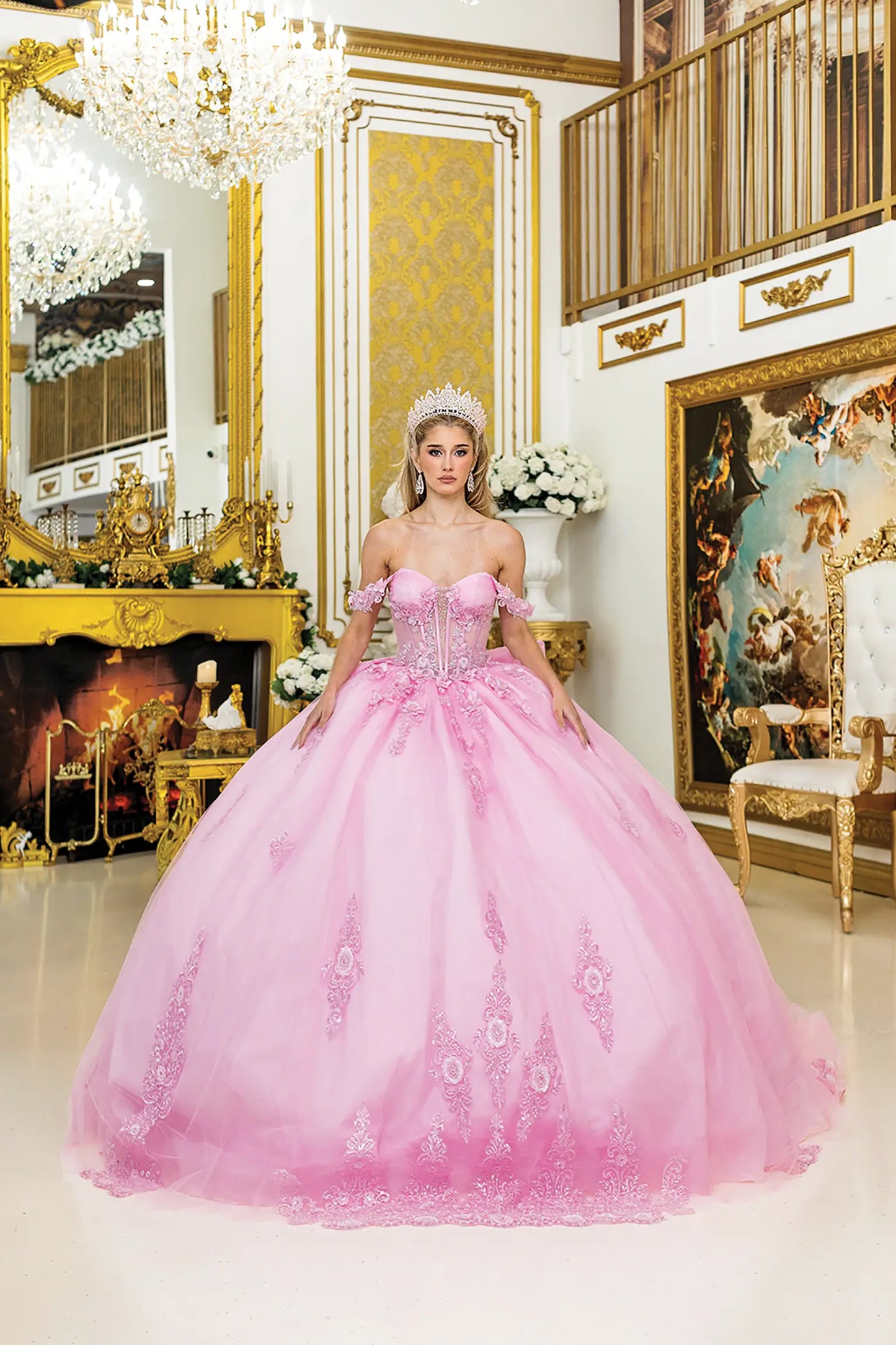 Woman standing front-facing in pink quinceañera ball gown with off-shoulder floral appliqué sleeves, sheer corset bodice, embroidered skirt accents, and voluminous elegant silhouette