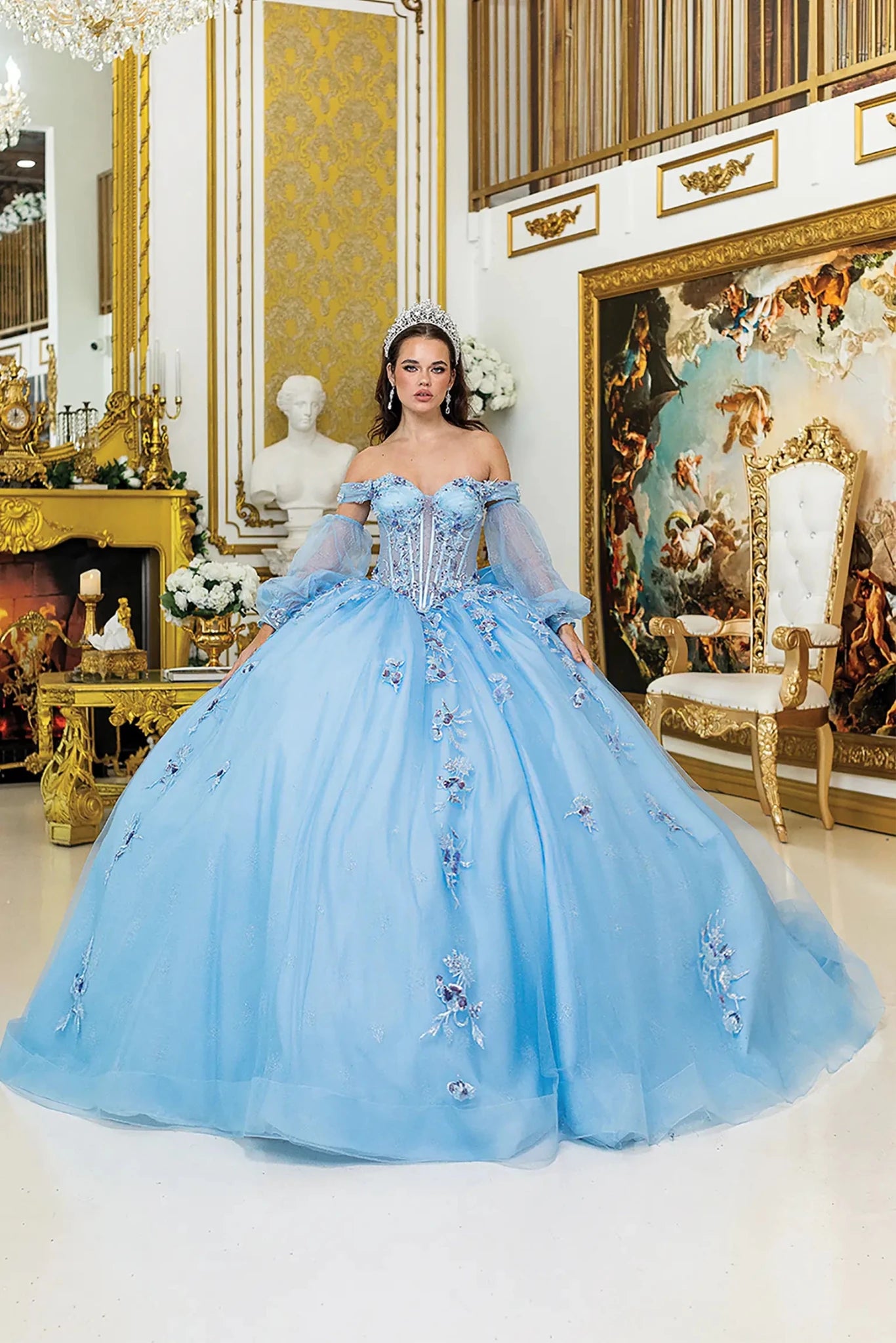Woman standing front-facing in Bahama blue quinceañera ball gown with sheer off-shoulder sleeves, structured corset bodice, floral appliqués, and voluminous glitter tulle skirt