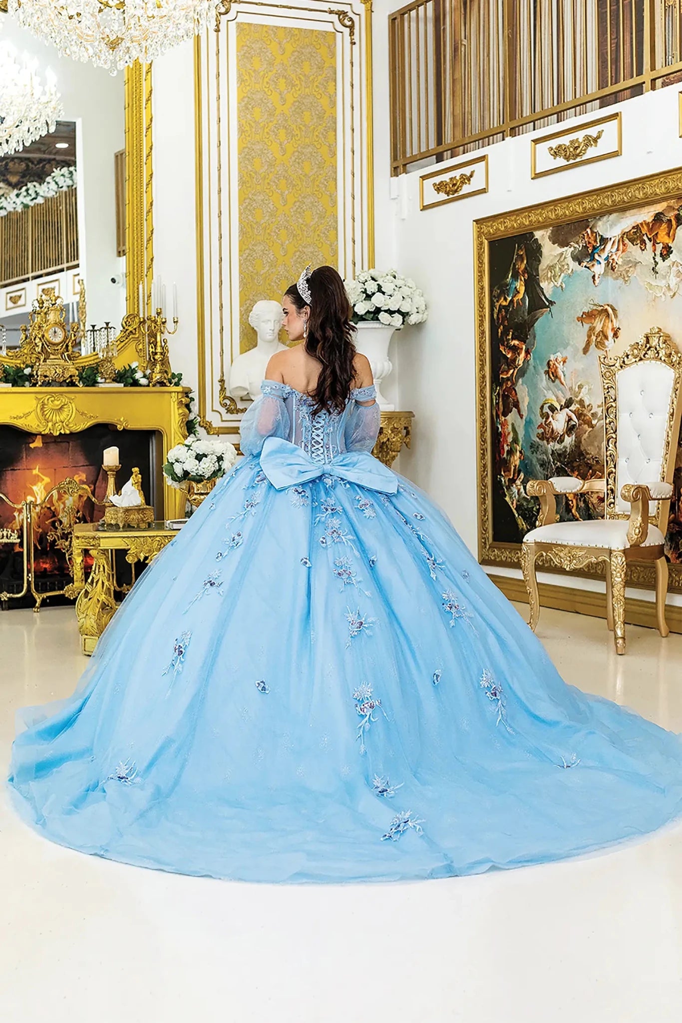 Woman standing back-facing in Bahama blue quinceañera ball gown with lace-up corset bodice, sheer off-shoulder sleeves, floral appliqués, and flowing glitter tulle skirt with train