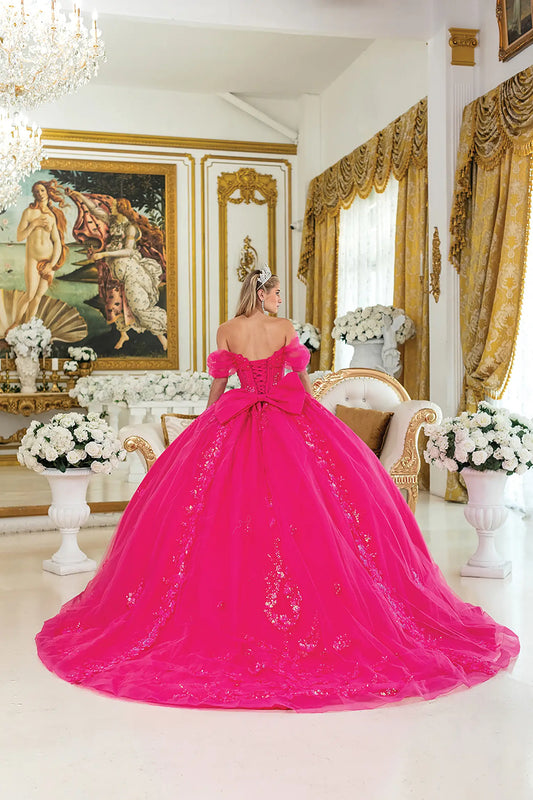 Woman standing back-facing in fuchsia quinceañera ball gown with lace-up corset bodice, off-shoulder sleeves, floral embroidery, glitter accents, and flowing tulle skirt with train