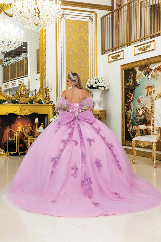 Woman standing back-facing in lilac-blush quinceañera ball gown with lace-up corset bodice, off-shoulder sheer sleeves, floral embroidery, 3D appliqués, and flowing layered tulle skirt with train