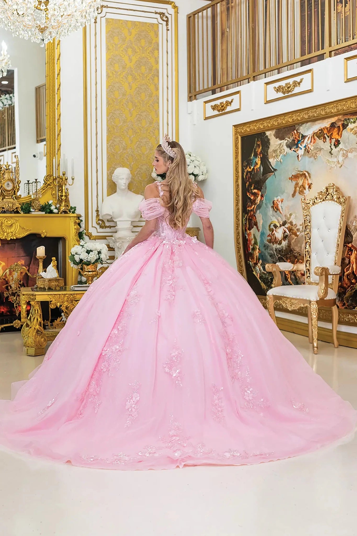 Woman standing back-facing in pink quinceañera ball gown with lace-up bodice, off-shoulder sleeves, floral appliqués, and flowing layered tulle skirt with train