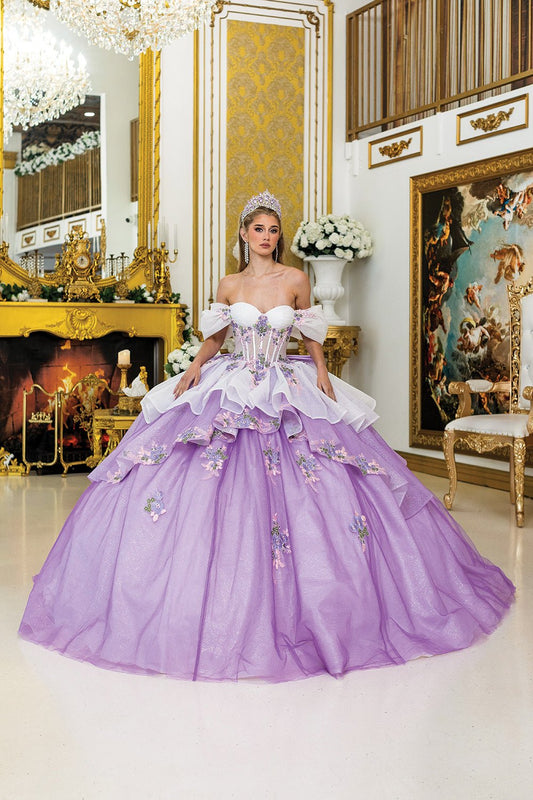 Woman standing front-facing in lilac-white quinceañera ball gown with off-shoulder sleeves, embroidered bodice, floral embellishments, and layered voluminous skirt