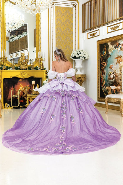 Woman standing back-facing in lilac-white quinceañera ball gown with lace-up embroidered bodice, floral embellishments, and flowing layered skirt with train