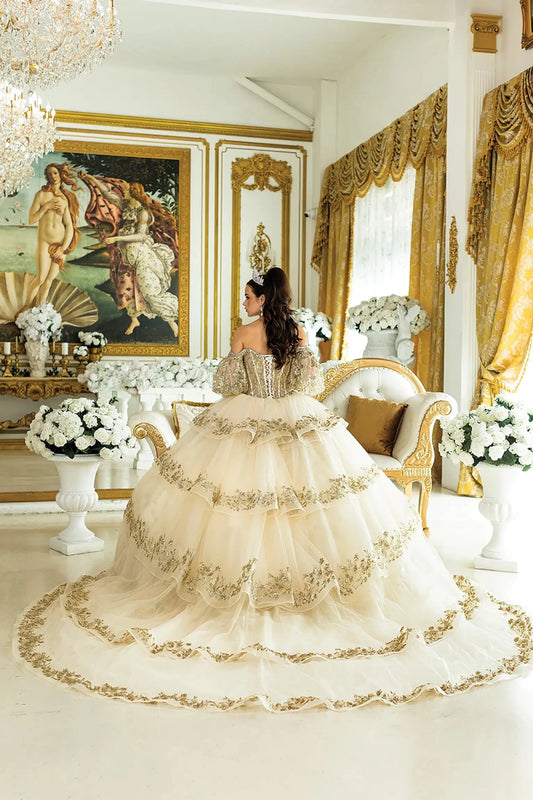 Woman standing back-facing in champagne quinceañera ball gown with off-shoulder sleeves, lace-up embroidered bodice, glitter accents, and flowing full skirt with train