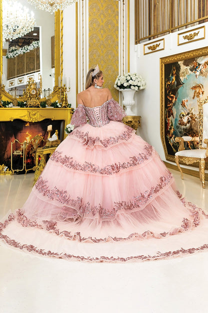 Woman standing back-facing in rose gold quinceañera ball gown with lace-up embroidered bodice, off-shoulder sleeves, glitter accents, and flowing layered skirt with train

