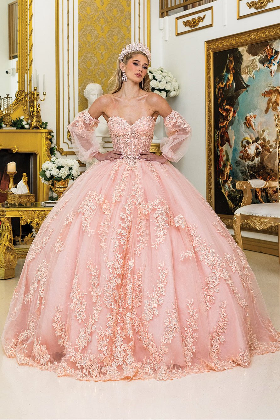 Woman standing front-facing in blush quinceañera ball gown with detachable sheer sleeves, floral appliqué detailing, corset bodice, and layered voluminous skirt
