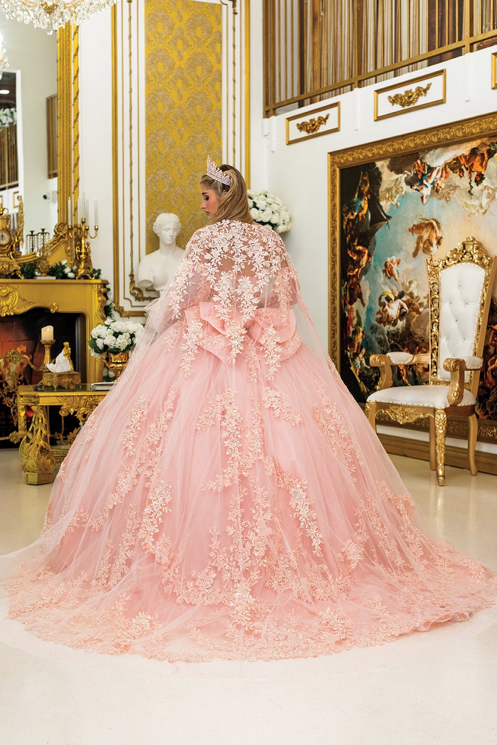 Woman standing back-facing in blush quinceañera ball gown with detachable sheer sleeves, corset bodice, floral appliqué accents, and shimmering layered skirt