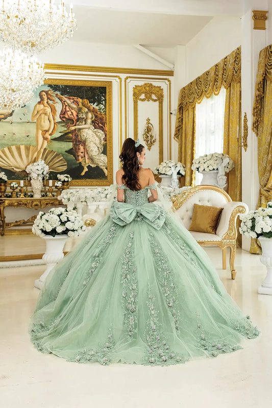 Woman standing back-facing in sage quinceañera ball gown with lace-up corset bodice, off-shoulder floral appliqués, and flowing sparkling tulle skirt with train