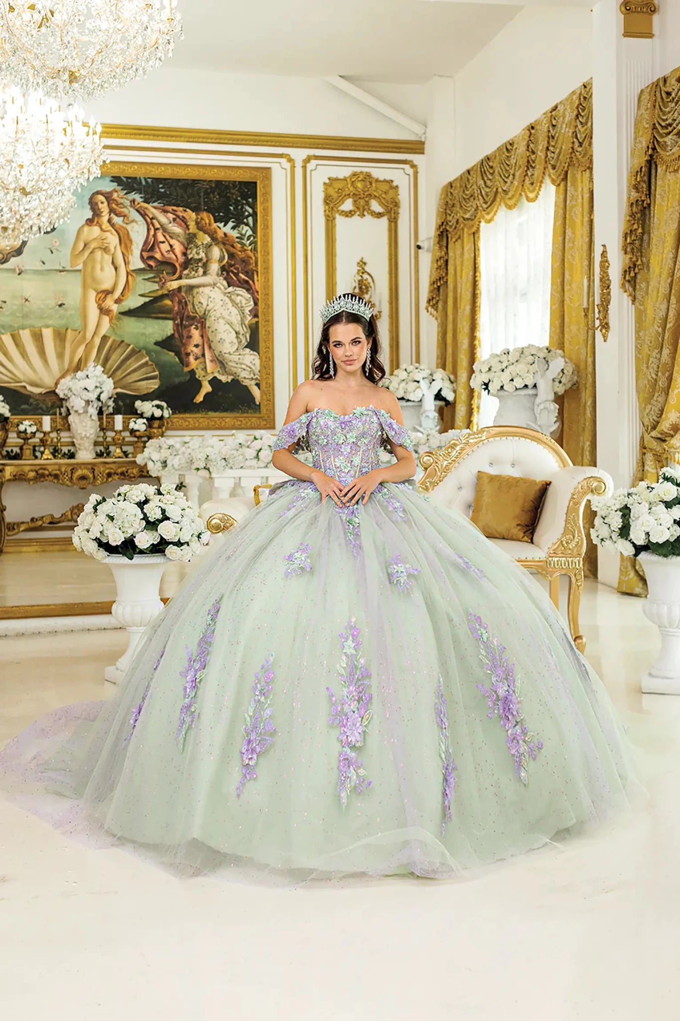 Woman standing front-facing in lilac-sage quinceañera ball gown with off-shoulder floral appliqués, corset bodice, glittered tulle skirt, and layered detailing
