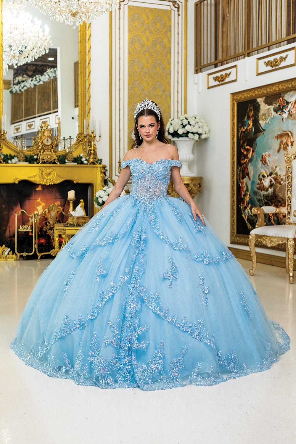Woman standing front-facing in blue quinceañera ball gown with rhinestone bodice, off-shoulder sleeves, embroidered detailing, and multi-tiered illusion skirt