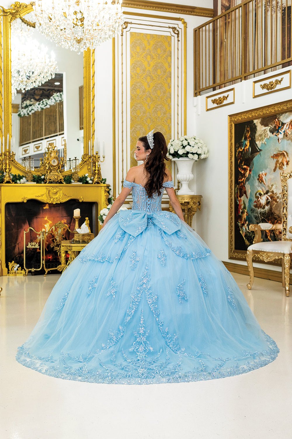 Woman standing back-facing in blue quinceañera ball gown with lace-up rhinestone bodice, off-shoulder sleeves, embroidered detailing, and flowing multi-tiered illusion skirt with train