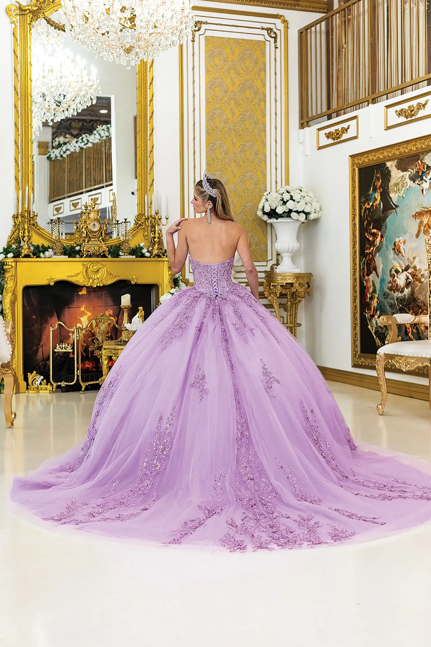 Back view of lilac quinceañera ball gown featuring a lace-up corset back, flowing tulle skirt with lace detailing, and an elegant sweep train in a luxurious gold room.