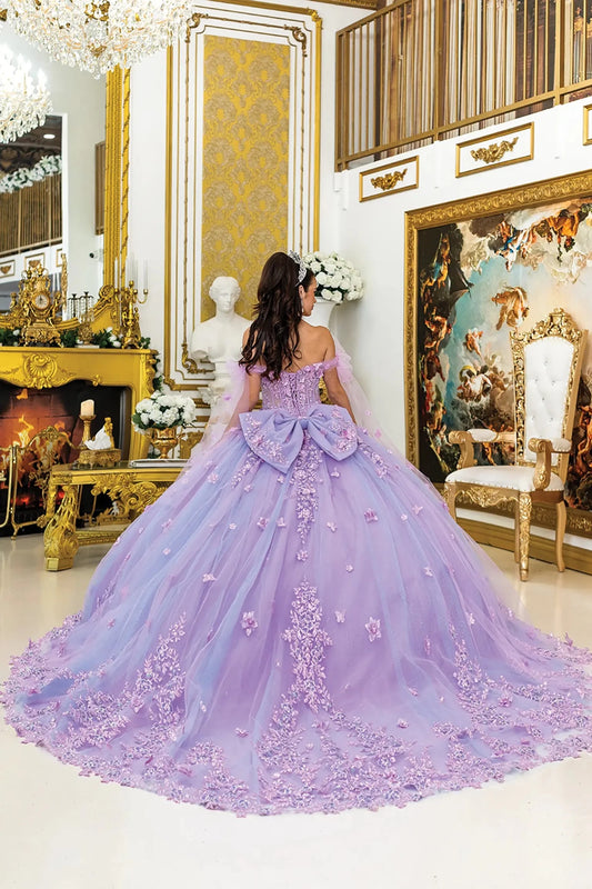 Woman standing back-facing in lilac quinceañera ball gown with lace-up corset bodice, off-shoulder sleeves, floral appliqués, and flowing layered glitter tulle skirt with train