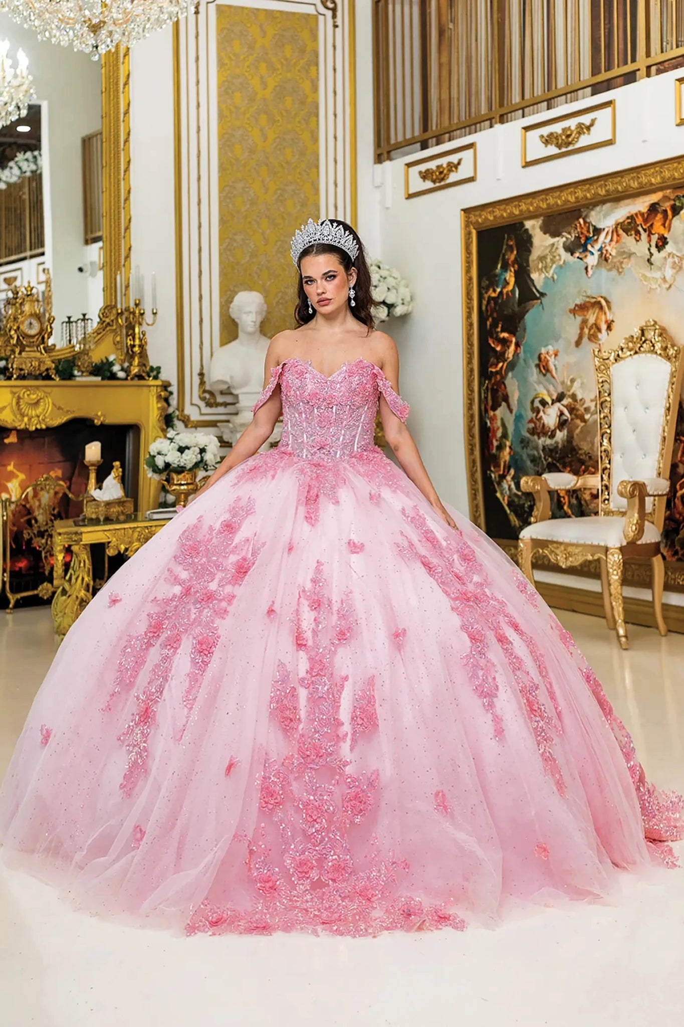 Woman standing front-facing in pink quinceañera ball gown with off-shoulder sleeves, fitted corset bodice, floral embroidery, and voluminous sparkling tulle skirt