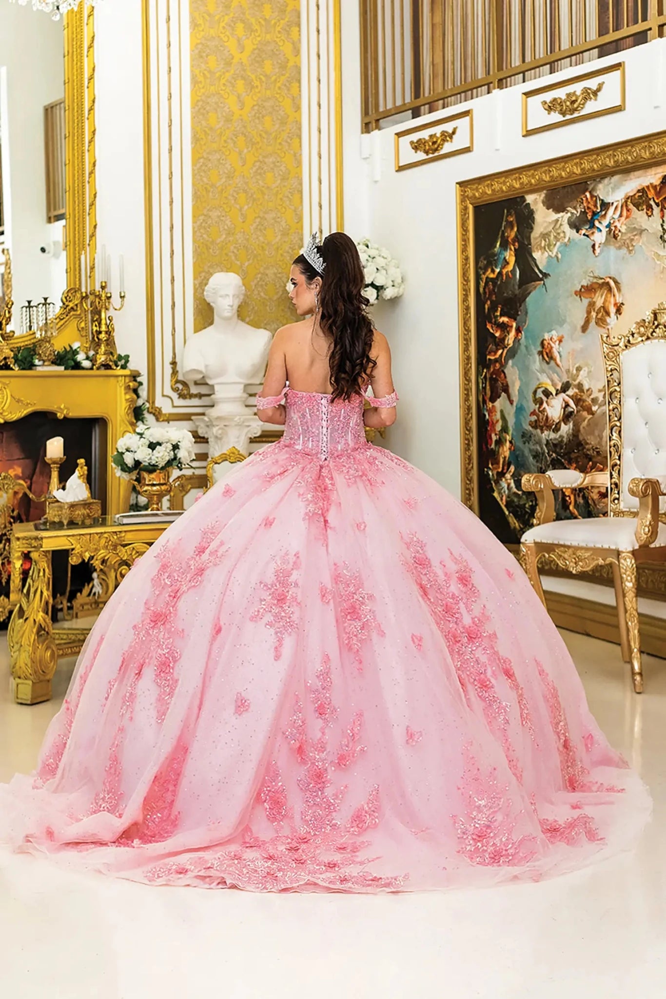 Woman standing back-facing in pink quinceañera ball gown with lace-up corset bodice, off-shoulder sleeves, floral embroidery, and flowing sparkling tulle skirt with train