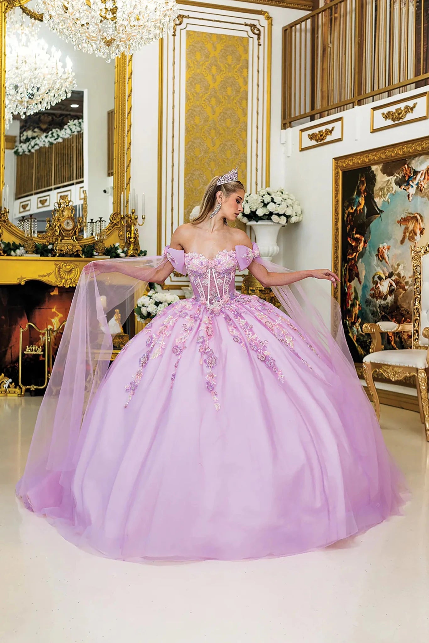 Woman standing front-facing in lilac quinceañera ball gown with off-shoulder sleeves, corset bodice, floral embroidery, glitter tulle overlay, and detachable sheer cape