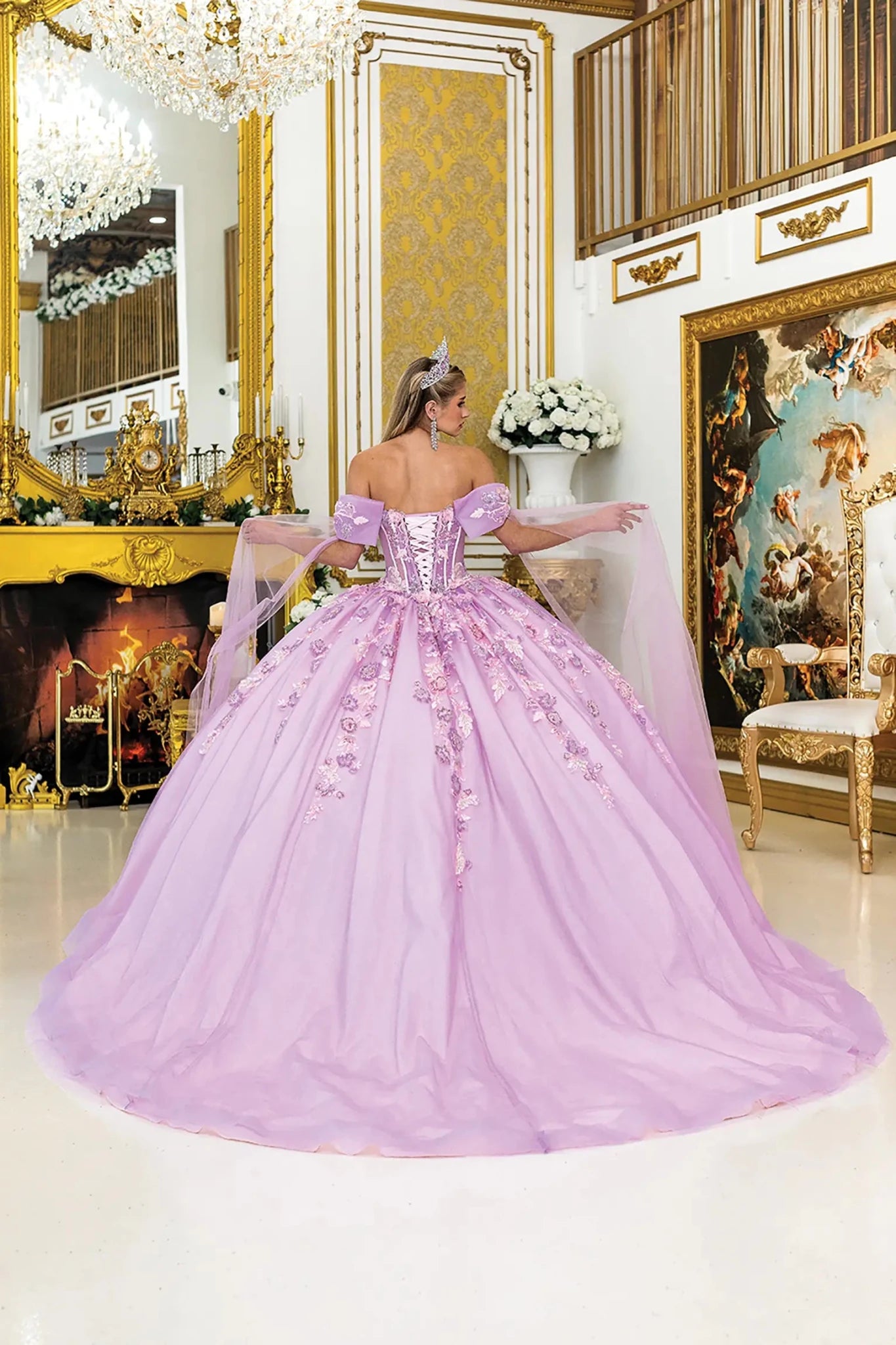 Woman standing back-facing in lilac quinceañera ball gown with off-shoulder sleeves, lace-up corset bodice, floral embroidery, glitter tulle overlay, and detachable sheer cape trailing behind