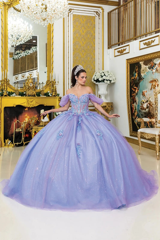 Woman standing front-facing in lilac-bahama blue quinceañera ball gown with corset bodice, off-shoulder puff sleeves, 3D floral embellishments, sheer panels, and voluminous glitter tulle skirt