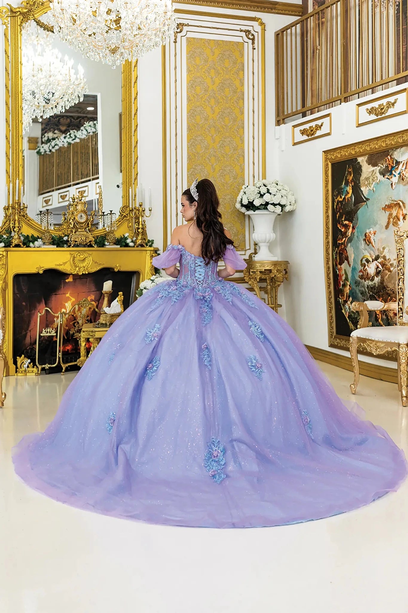 Woman standing back-facing in lilac-bahama blue quinceañera ball gown with lace-up corset bodice, off-shoulder puff sleeves, 3D floral embellishments, sheer panels, and flowing glitter tulle skirt with train